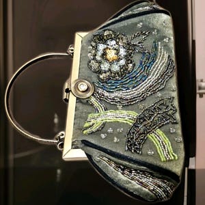 May include: A grey clutch bag with a silver handle and clasp. The bag is decorated with a floral design and beaded patterns in silver, blue, green, and gold. The bag has a rectangular shape.
