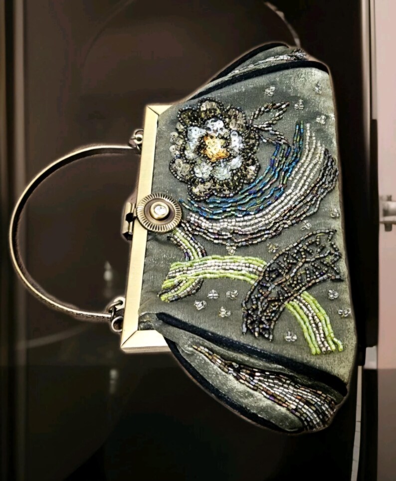 May include: A grey clutch bag with a silver handle and clasp. The bag is decorated with a floral design and beaded patterns in silver, blue, green, and gold. The bag has a rectangular shape.