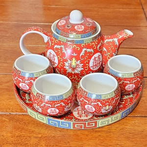 May include: A red and white ceramic tea set with a teapot, four cups, and a serving tray. The set features a floral and geometric design with Chinese characters. The teapot has a white handle and a white lid. The tray is round.