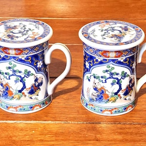 Può includere: Due tazze in ceramica con coperchi, con un design di ispirazione asiatica. Le tazze hanno manici bianchi e sono decorate con motivi blu, rossi e oro, tra cui figure e motivi floreali. I coperchi rispecchiano il design.