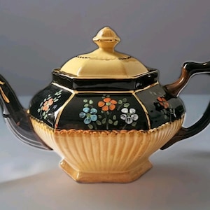 May include: A vintage teapot with a black and gold design. The teapot features a yellow lid and base, with a black body adorned with colorful floral patterns. The handle and spout are black with gold accents. The teapot is approximately 10 inches long.