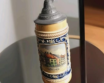 May include: A ceramic beer stein with a pewter lid. The stein features a painted scene of buildings with the text "MÜNCHEN" and "DEUTSCHLAND".