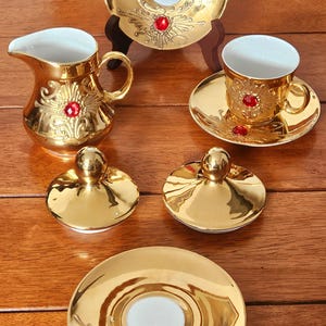 May include: A gold tea set featuring a creamer, teacup, saucer, and lids, all adorned with floral patterns and red jewel embellishments. The set is displayed on a wooden surface, showcasing its elegant design.