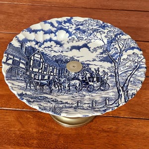 Può includere: Una ciotola rotonda in ceramica su piedistallo con un motivo blu e bianco che raffigura una carrozza trainata da cavalli davanti a un edificio a graticcio. La ciotola presenta un bordo smerlato e un punto di fissaggio centrale in metallo.
