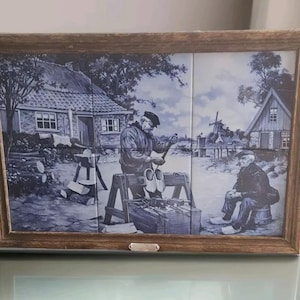 Könnte beinhalten: Gerahmtes blau-weißes Kachelkunstwerk, das eine ländliche Szene mit zwei Männern, einer Windmühle und einem Haus zeigt. Das Kunstwerk ist in einem Holzrahmen gefasst. Die Szene zeigt einen Mann, der an Holzschuhen arbeitet, und einen anderen Mann, der auf einem Fass sitzt.