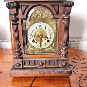 May include: A vintage wooden mantel clock with a gold-toned clock face and ornate detailing. The clock features Roman numerals and decorative accents. A pendulum and key are visible on the wooden surface in front of the clock.