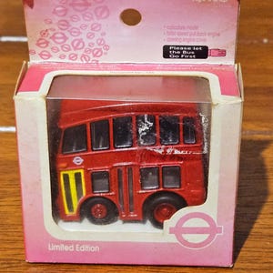 May include: A red toy double-decker bus in a pink box. The bus has black wheels and windows, with a yellow and black logo on the side. The box reads "Limited Edition" and "Age 3 & up". The text "Please let the Bus Go First" is also visible.