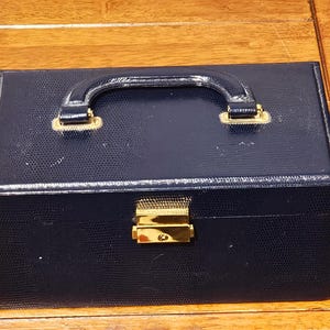 May include: A navy blue jewellery box with a textured, faux-leather exterior. It features a curved handle and gold-coloured metal accents on the handle and clasp. The box is rectangular and sits on a wooden surface.