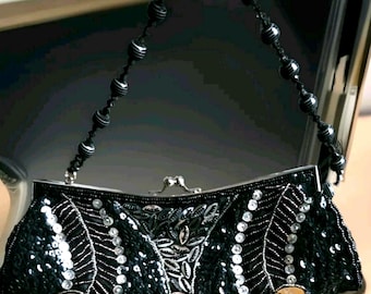 Op de afbeelding: Een zwarte clutch met ingewikkelde kralen- en paillettenversieringen in een bladvormig patroon. De tas heeft een zilverkleurige metalen knipsluiting en een zwart koordhengsel dat over de gehele lengte is versierd met ronde, geribbelde zwarte kralen.