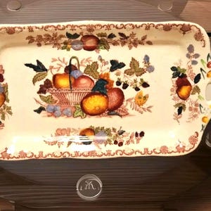 Op de afbeelding: Rechthoekige keramische serveerschaal met een crèmekleurige basis en een gedetailleerd fruitmandontwerp in het midden. De schaal heeft een bruine bloemenrand en handvatten aan beide uiteinden. De fruitmand bevat sinaasappels, appels en druiven.