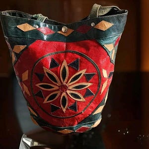 May include: A red, green and brown leather tote bag with a circular design featuring a flower motif. The bag has a top handle and a shoulder strap.