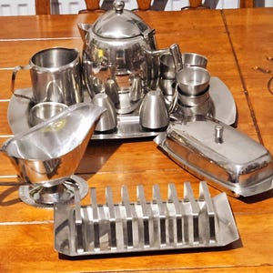 Könnte beinhalten: Ein komplettes Tee- und Frühstücks-Set aus Edelstahl. Das Set beinhaltet eine Teekanne, einen Sahnekännchen, eine Zuckerdose, Salz- und Pfefferstreuer, eine Butterdose und einen Toastständer. Die Artikel sind auf einem Holztisch angeordnet.