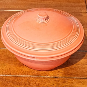 Puede incluir: Un cuenco de cerámica de color coral con tapa a juego. El cuenco y la tapa tienen un diseño acanalado. La tapa tiene un pequeño asa redonda. El cuenco está sobre una superficie de madera.