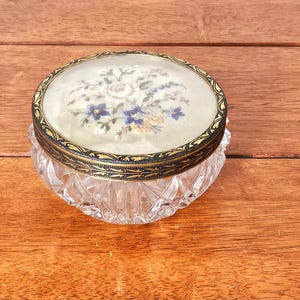 Puede incluir: Un frasco de tocador redondo de vidrio con base de cristal transparente tallado y tapa de metal. La tapa presenta un borde decorativo negro y dorado que rodea una incrustación de tela bordada con un ramo floral en tonos blanco, azul y amarillo.