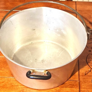 May include: A large, round, silver-colored aluminum cooking pot with a smooth interior and exterior. It features a single, arched metal bail handle attached to the sides and a small, black, horizontal handle mounted on the front rim.