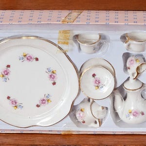 Puede incluir: Un juego de té de porcelana en miniatura en una caja rosa y blanca. El juego incluye un plato grande, tazas pequeñas, platillos, una tetera y una crema. Cada pieza está decorada con motivos florales rosas y azules y ribetes dorados.