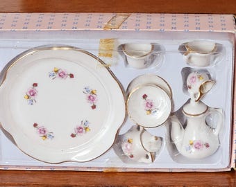 May include: A miniature porcelain tea set in a pink and white box. The set includes a large plate, small cups, saucers, a teapot, and a creamer. Each piece is decorated with pink and blue floral patterns and gold trim.