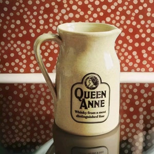 May include: A cream-coloured ceramic jug with a handle. The jug has a black and white label that reads "Queen Anne Whisky from a most distinguished line".