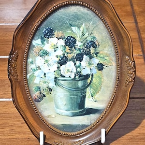 May include: An oval framed print depicting a still life of blackberries and white flowers in a metal bucket. The brown frame has decorative detailing. The artwork is displayed on a white stand.
