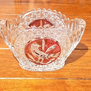 Puede incluir: Un cuenco de cristal tallado transparente con un diseño rojo con temática de pájaros. El cuenco tiene un borde festoneado y un patrón geométrico. El diseño del pájaro tiene forma ovalada, con un pájaro y follaje.