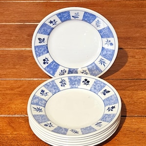 Puede incluir: Una pila de platos de cerámica blanca con un diseño floral azul y blanco. Los platos tienen un borde azul a cuadros con motivos florales y de hojas. Los platos están apilados sobre una superficie de madera.