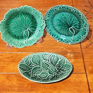 Op de afbeelding: Drie groene keramische borden in de vorm van bladeren. Twee zijn rond met een groot bladerontwerp en de derde is ovaal met een gelaagd bladerpatroon. De borden staan op een houten oppervlak.