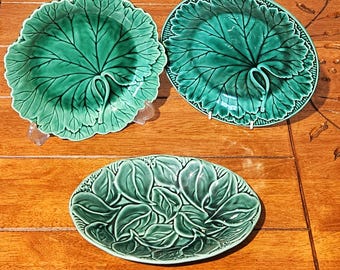 Puede incluir: Tres platos verdes de cerámica con forma de hoja. Dos son redondos con un diseño de hoja grande, y el tercero es ovalado con un patrón de hojas en capas. Los platos están sobre una superficie de madera.