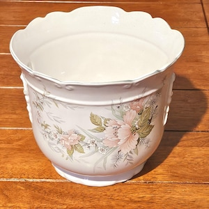 Könnte beinhalten: Ein weißer Keramikübertopf mit gewelltem Rand und einem Blumenmuster aus zartrosa Pfingstrosen, grünen Blättern und kleinen weißen Blüten. Das zylindrische Gefäß hat eine glatte, glänzende Oberfläche und einen leicht ausgestellten Rand.