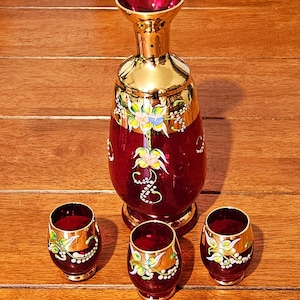 Könnte beinhalten: Ein rubinrotes Glasdekanter und drei passende Schnapsgläser. Das Set hat goldene Ränder und Blumendesigns in Grün, Gelb und Blau. Die Karaffe ist hoch mit breitem Boden, und die Schnapsgläser sind klein und abgerundet.