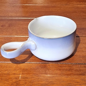 May include: A light blue ceramic soup bowl with a distinctive loop handle. The bowl has a white interior and a textured finish. The handle is designed for easy gripping. The bowl is displayed on a wooden surface.