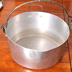 Puede incluir: Una olla grande de aluminio con un asa curva y un asa más pequeña en el lateral. La olla tiene una abertura ancha y un fondo redondeado. La olla está sobre una superficie de madera.