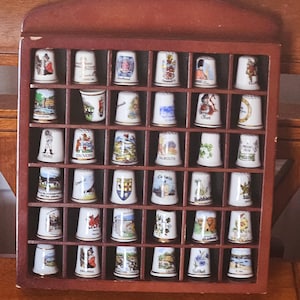 Puede incluir: Un expositor de madera con 30 dedales decorativos. Cada dedal es blanco y presenta ilustraciones coloridas de lugares y figuras. El expositor es de color marrón rojizo oscuro.