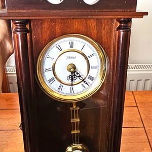 May include: A dark brown wooden mantel clock with a pediment top and a central finial. The clock face is white with black Roman numerals and the text Highlands. It features a gold-toned bezel, black ornate hands, and a gold-toned pendulum.