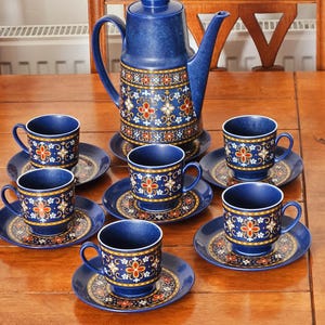 Puede incluir: Un juego de café vintage de cerámica azul que incluye una cafetera y seis tazas con platillos. El juego presenta un patrón floral en rojo, amarillo y blanco. La cafetera es alta con un pico y asa curvados. Las tazas y platillos están sobre una mesa de madera.