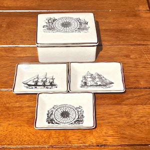 May include: A white ceramic box and four matching rectangular dishes with nautical designs. The box lid and one dish feature a compass rose design. The other dishes depict sailing ships. All have a brown trim.