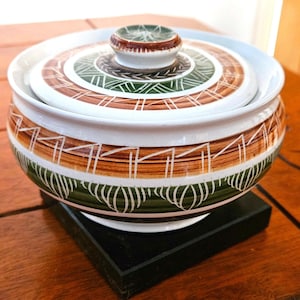 May include: A ceramic bowl with a lid, decorated with brown, green, and white geometric patterns. The bowl rests on a black square base. The lid has a small, round handle.