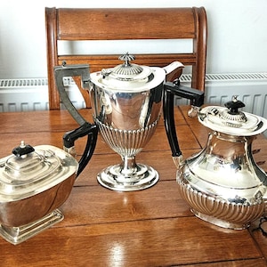 May include: Three antique silver tea sets are arranged on a wooden table. The tea sets include a teapot, a sugar bowl, and a creamer. The teapot has a curved spout and a black handle. The sugar bowl is rectangular with a black handle.