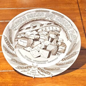 May include: A white ceramic plate with a brown illustration of bread, cheese, and a bottle. The plate is labeled "BRITISH BREAD AND CHEESE PLATE" with wheat stalks around the edge. Cheese varieties listed include Stilton and Cheddar.
