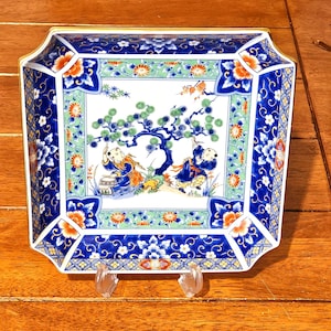 May include: A rectangular ceramic serving dish with a detailed, colorful design. The dish features a central scene of children under a tree, surrounded by floral and geometric patterns in blue, orange, and green. The edges are trimmed with gold.