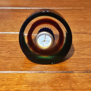 Può includere: Un orologio da scrivania rotondo in vetro con quadrante. L'orologio è racchiuso in un cilindro di vetro trasparente con un anello esterno verde scuro, un anello interno marrone-rossastro e un quadrante argentato con la scritta "Calthness".