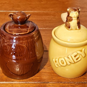 May include: A brown ceramic barrel-shaped container with a lid topped by a small teapot, embossed with the text TEA TIME, alongside a yellow ceramic jar with a lid featuring a bear figurine, embossed with the word HONEY in raised letters.