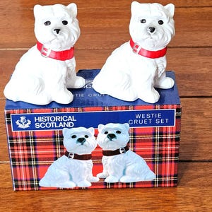 May include: Two white West Highland Terrier figurines with red collars. They sit on a box with a red and black tartan pattern. The box is labelled "Historical Scotland Westie Cruet Set". The figurines have black eyes and noses.