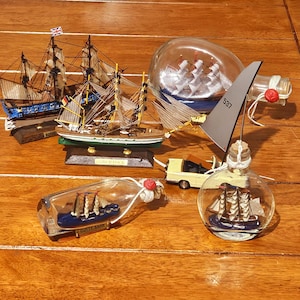 Puede incluir: Una colección de modelos de barcos en miniatura, incluyendo barcos en botellas y veleros independientes. Los barcos presentan detalles intrincados, con velas, mástiles y banderas. La escena está ambientada sobre una superficie de madera, mostrando el tema náutico.