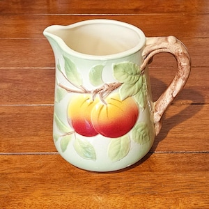 May include: A ceramic pitcher featuring a light green background with a raised, three-dimensional design of two red and yellow peaches on a branch with green leaves. The handle is textured to resemble a brown tree branch with a curved spout.