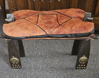 May include: A handcrafted wooden bench with a brown leather seat. The seat is intricately patterned with black stitching and a black border. The legs are dark brown with brass accents and rivets, creating a rustic aesthetic.