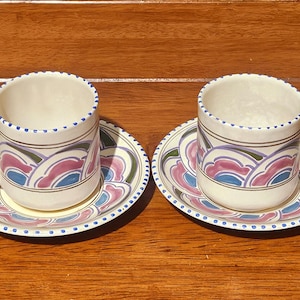 May include: Two matching ceramic teacups and saucers. The cups and saucers are white with a repeating pattern of pink, blue, and green abstract shapes. The rims are decorated with blue dots. The handles are white with a blue accent.