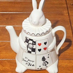May include: White ceramic teapot with a rabbit head lid and a ruffled collar. The teapot features illustrations of playing cards, a pocket watch, and Alice in Wonderland, with the text "It's Always Time for Tea."