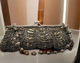 Puede incluir: Un bolso de mano de cuentas con cierre de plata y una correa de cadena. El bolso está hecho de cuentas negras y doradas y tiene un diseño festoneado.