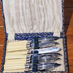 May include: An antique cutlery set in a blue and cream lined box. The set includes knives, forks, and spoons with cream-colored handles and silver blades. The box has a blue patterned exterior and a cream-colored interior.