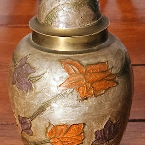 May include: A decorative, lidded jar with a metallic finish. The jar has a brass rim and a brass-coloured finial. The body is adorned with painted leaves in shades of orange, purple, and green, creating an autumnal theme.